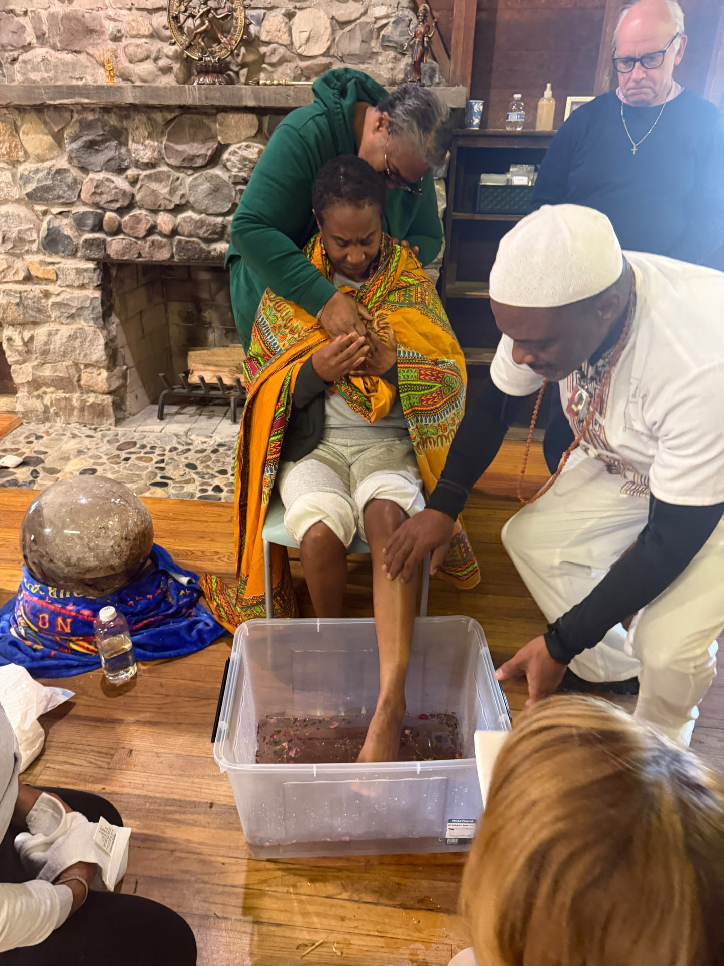 An elder wraps a participant in golden cloth by the stone fireplace
